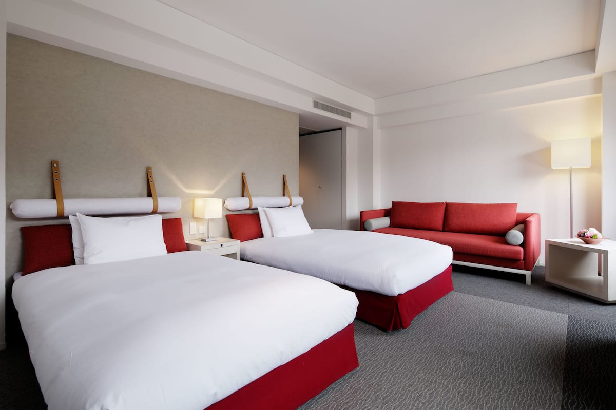 Habitación de hotel con dos camas, un sofá rojo, una mesita con una lámpara y un estante en la pared.