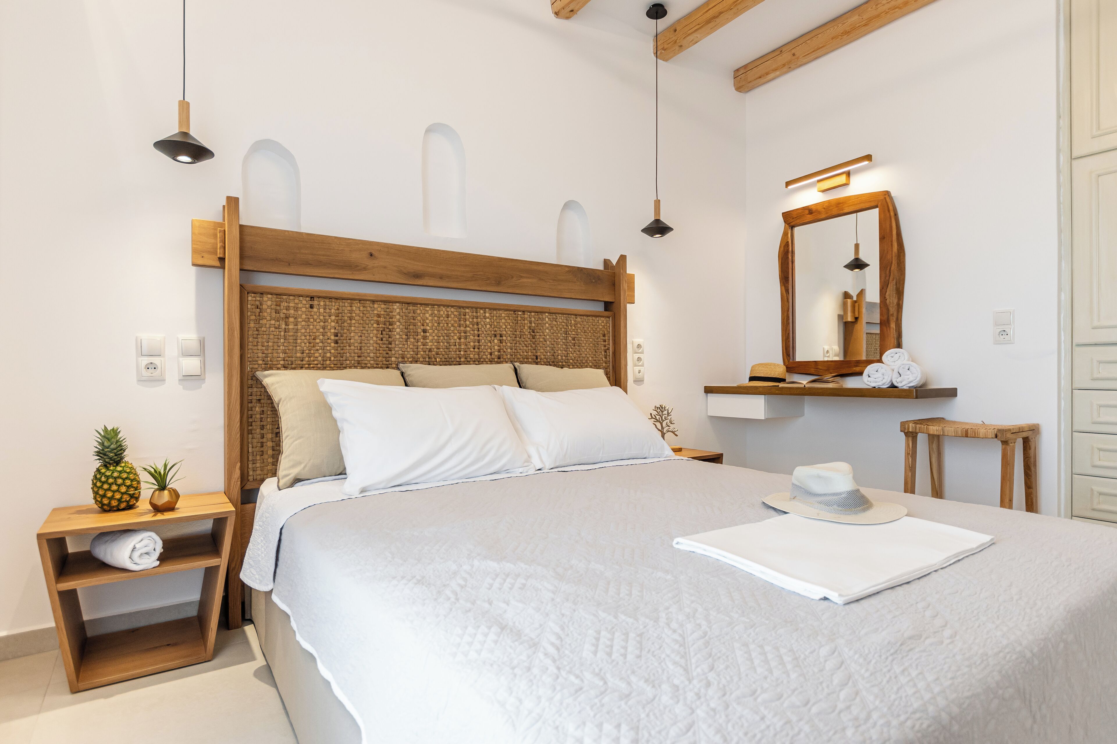 A neatly arranged bedroom with a wooden headboard, a bedside table with a rolled towel, a small plant, and a hat on the bed.