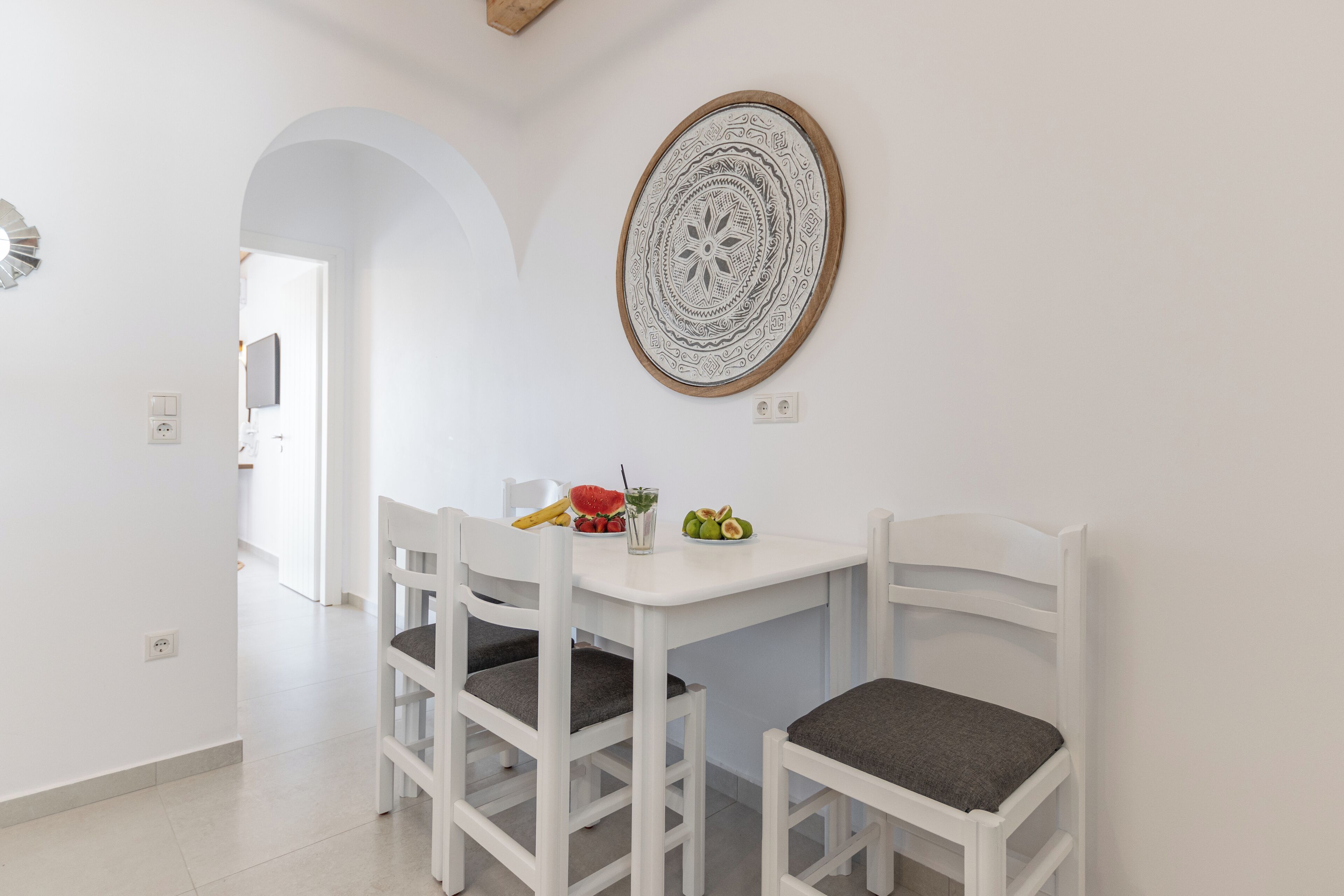 A dining area with a white table and four white chairs, a decorative wall piece, and a fruit bowl with various fruits.