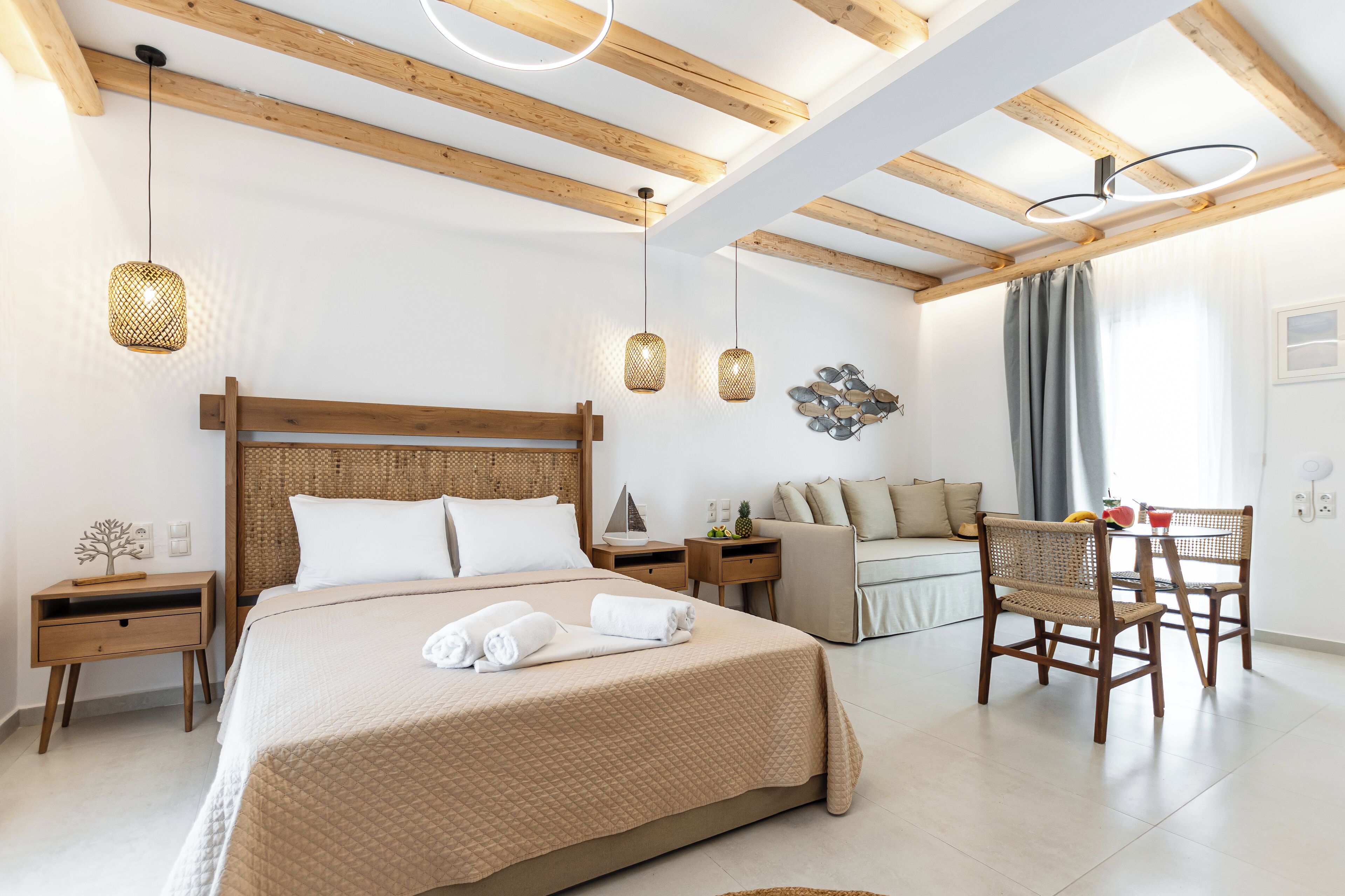 A modern hotel room with a large bed, wooden ceiling beams, a dining area with a table and chairs, and a small side table with a plant.