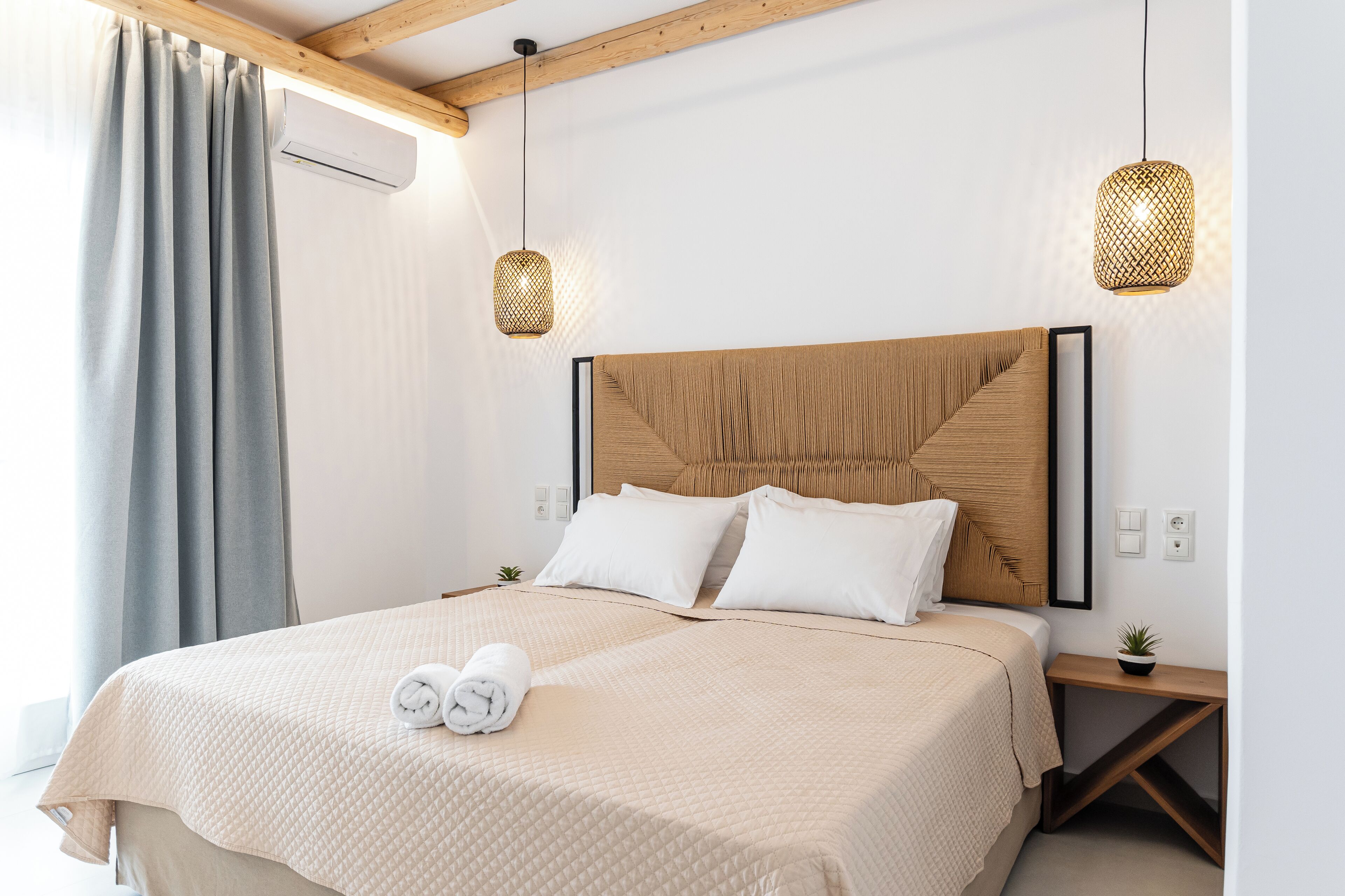 A neatly made bed with a beige quilt, two white pillows, and a rolled towel on the side. There are two bedside tables, one with a small plant. The room has wooden beams on the ceiling and two hanging lights.