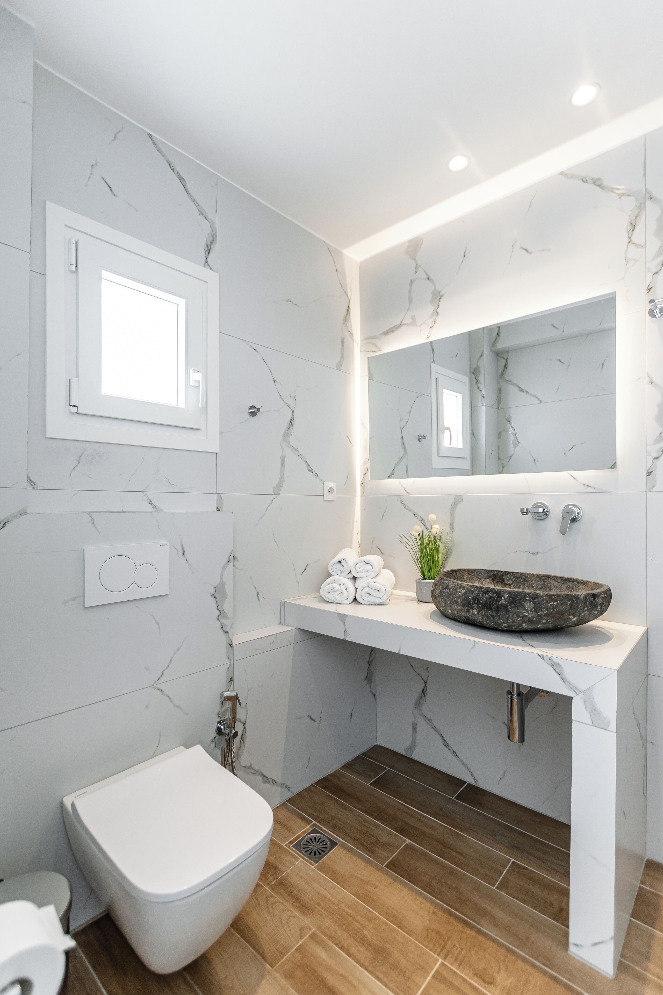 A modern bathroom with a marble backsplash, a white sink with a stone basin, a large mirror, and a toilet.