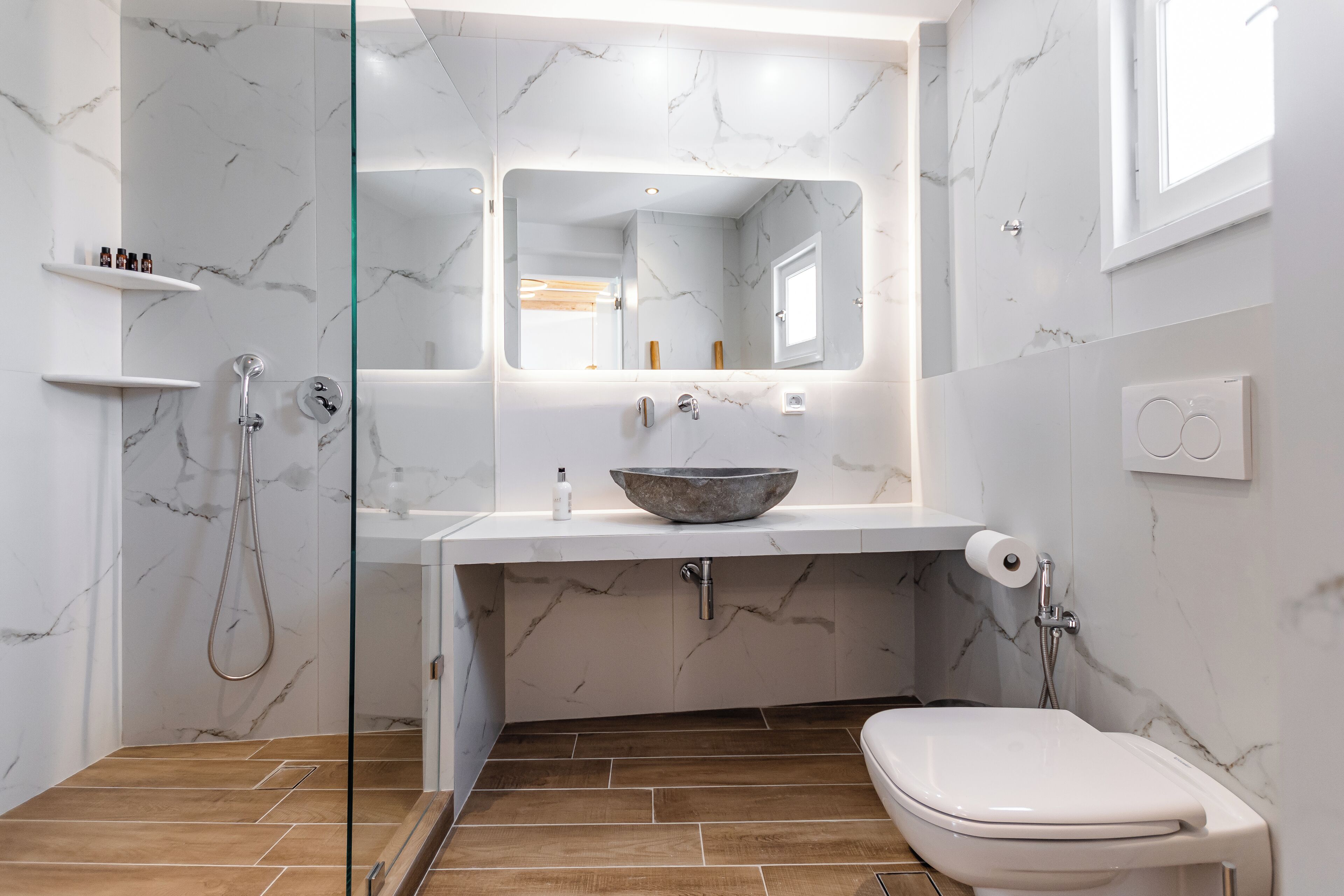 A modern bathroom with a glass shower enclosure, a marble-tiled vanity, a white toilet, and a large mirror.