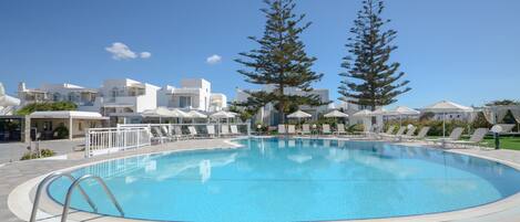 Una piscina con sillones de playa y sombrillas, rodeada de edificios blancos y altos árboles.
