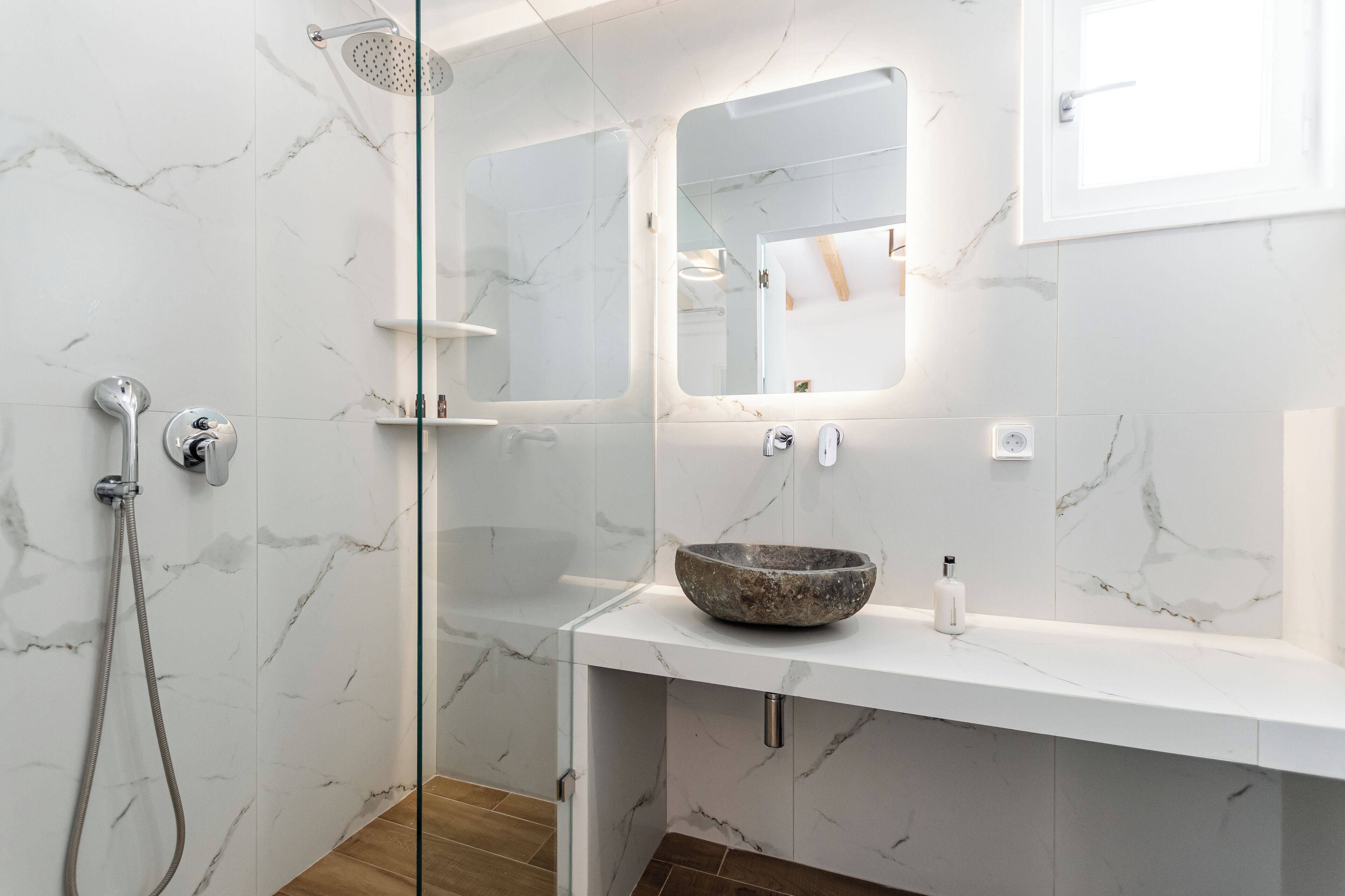 Ein modernes Badezimmer mit einer gläsernen Duschkabine, einer mit Marmor belegten Wand, einem weißen Waschbecken mit Natursteinbecken und einem Spiegel über dem Waschbecken.
