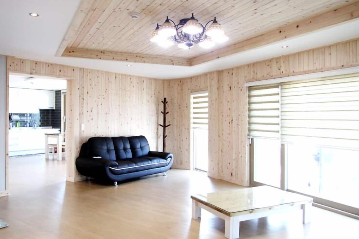 Un vaste salon aux murs lambrissés, avec un canapé en cuir noir, une table basse en verre et un lustre.