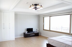 Un dormitorio con cama, televisor sobre un soporte negro, armario y un amplio ventanal con vistas al balcón.