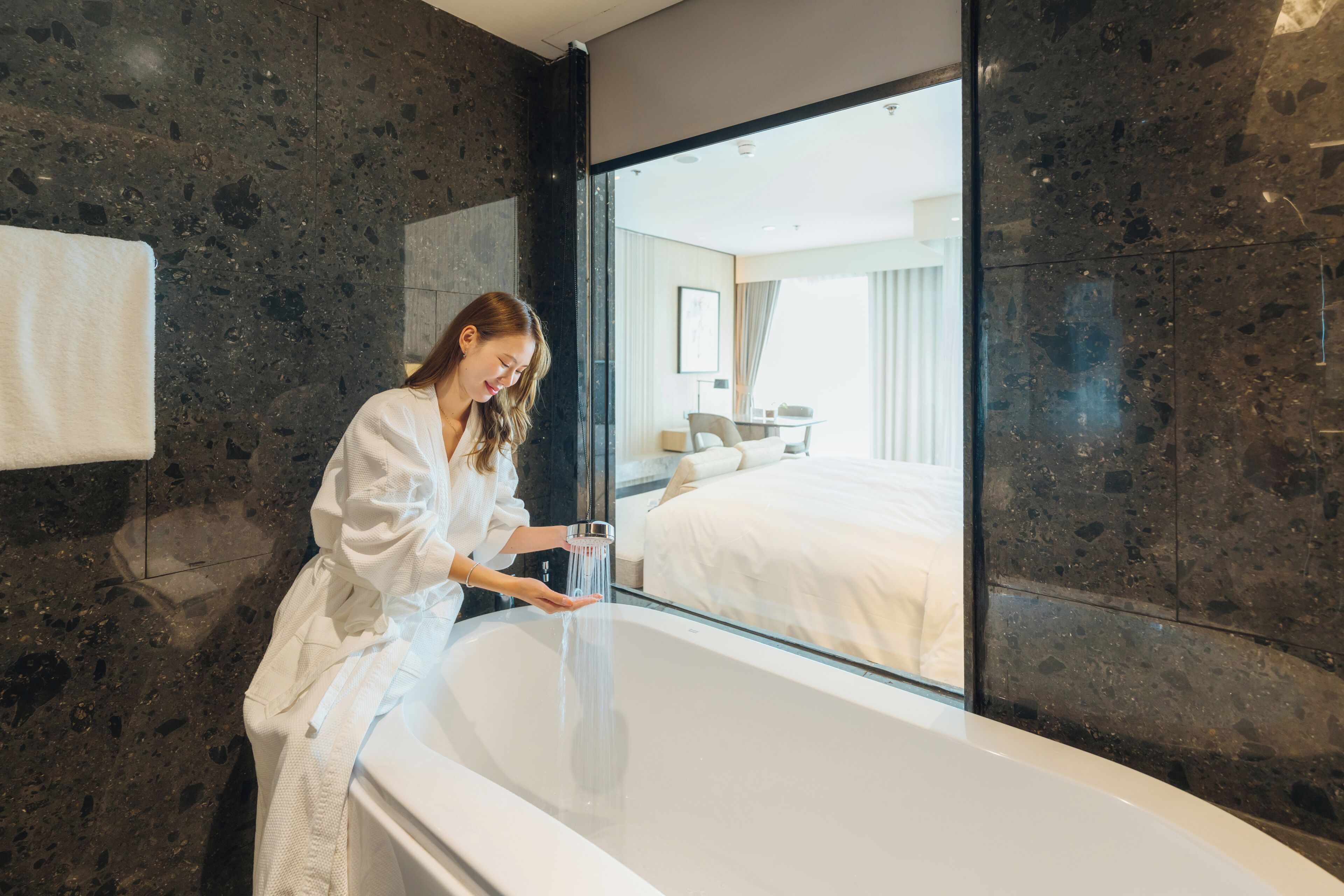 A modern bathroom with a large mirror, a bathtub, and a visible bedroom with a bed and curtains.