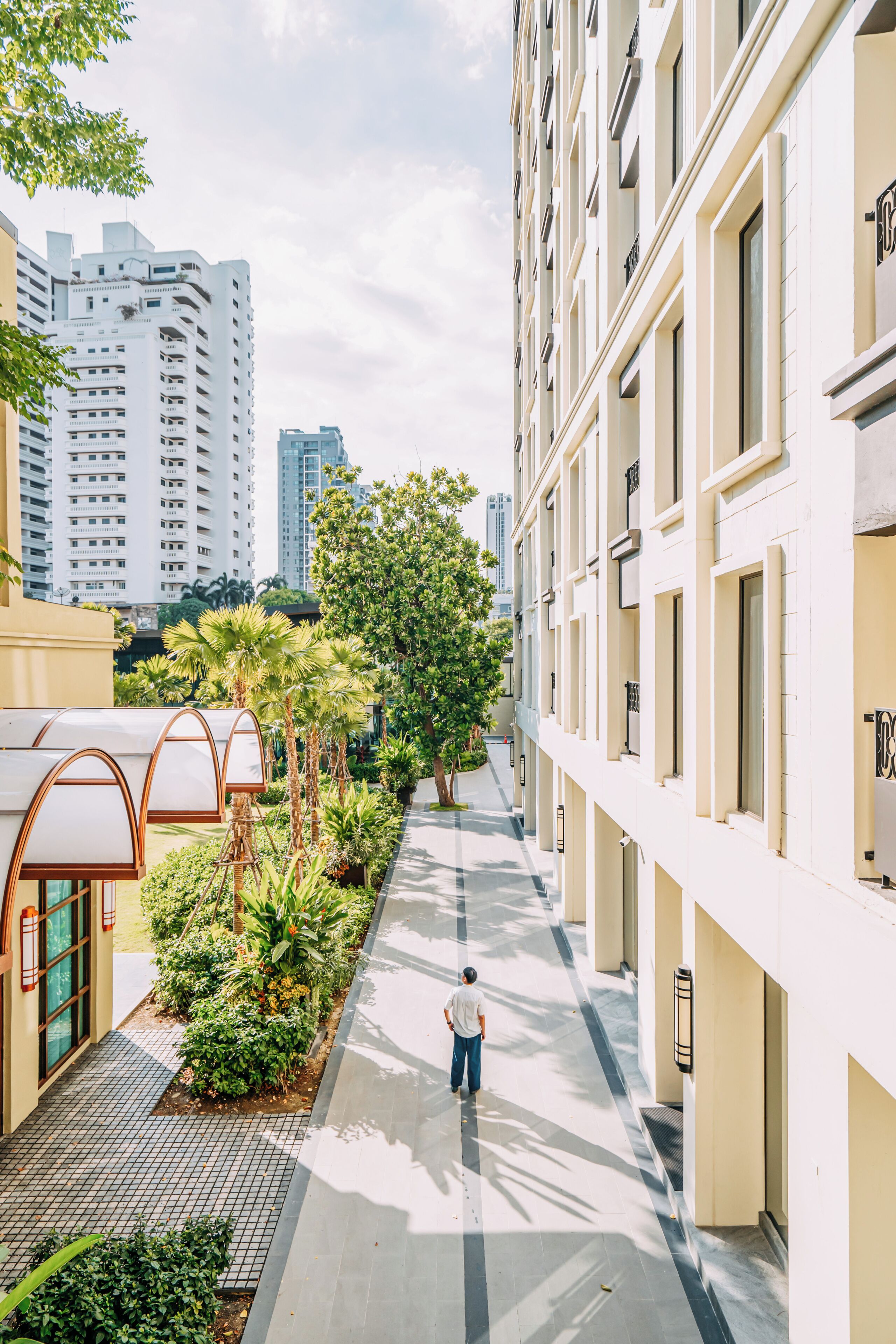 A corridor with a person walking, surrounded by greenery and buildings.