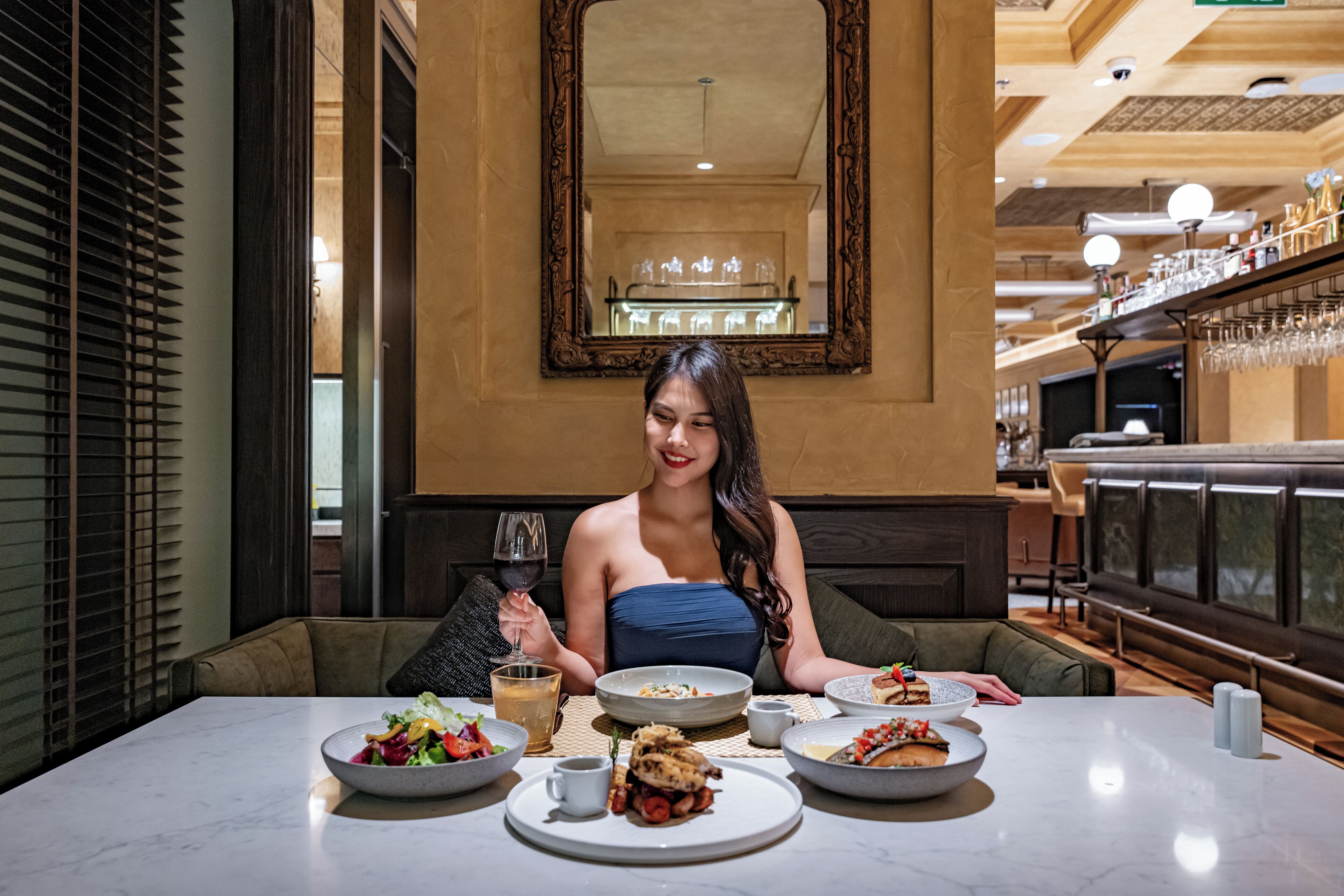 A woman seated at a table with various dishes, including a salad, a plate with a dessert, and a glass of wine.