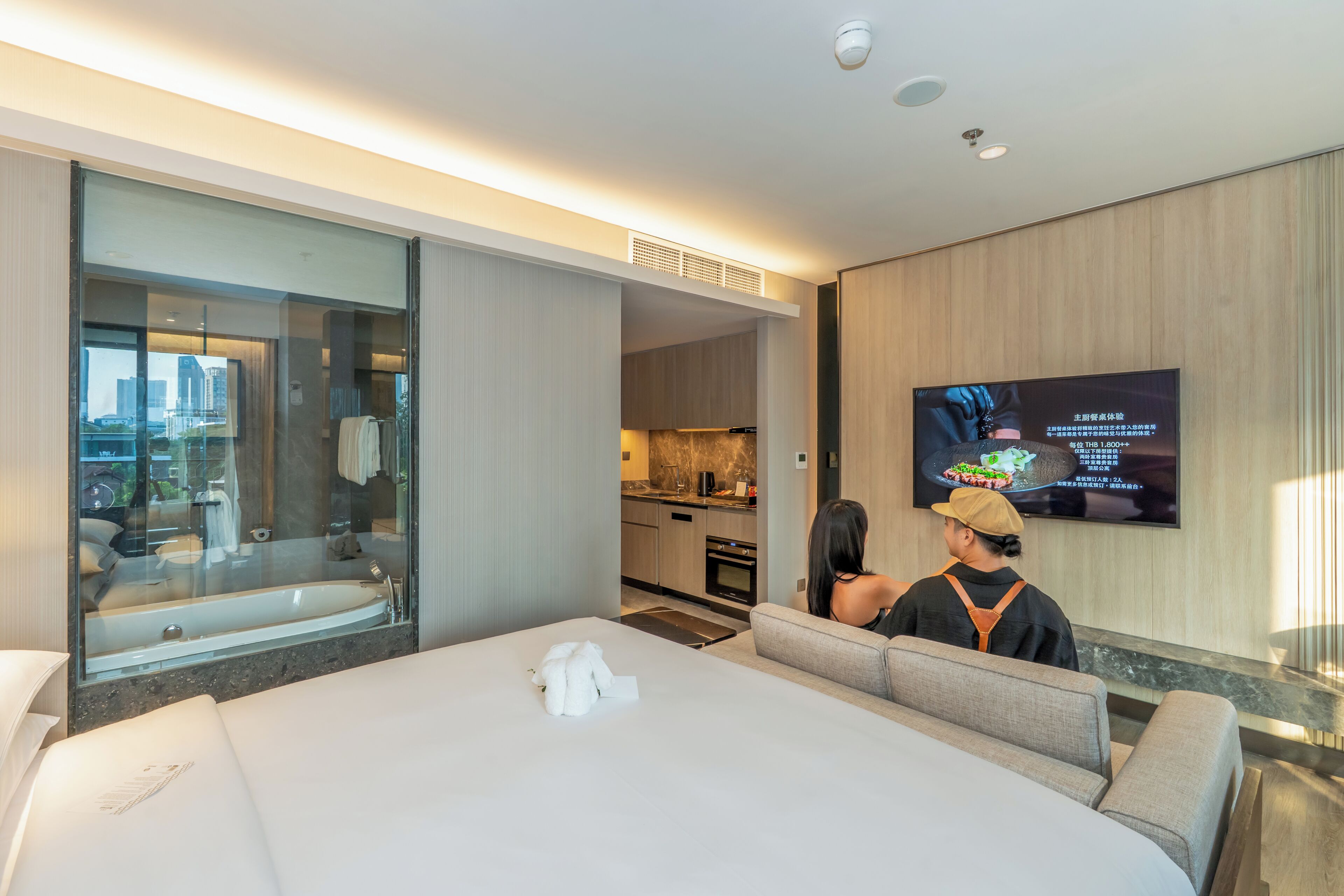 A hotel room with a large bed, a TV on the wall, a bathroom with a bathtub, and a view of the city.