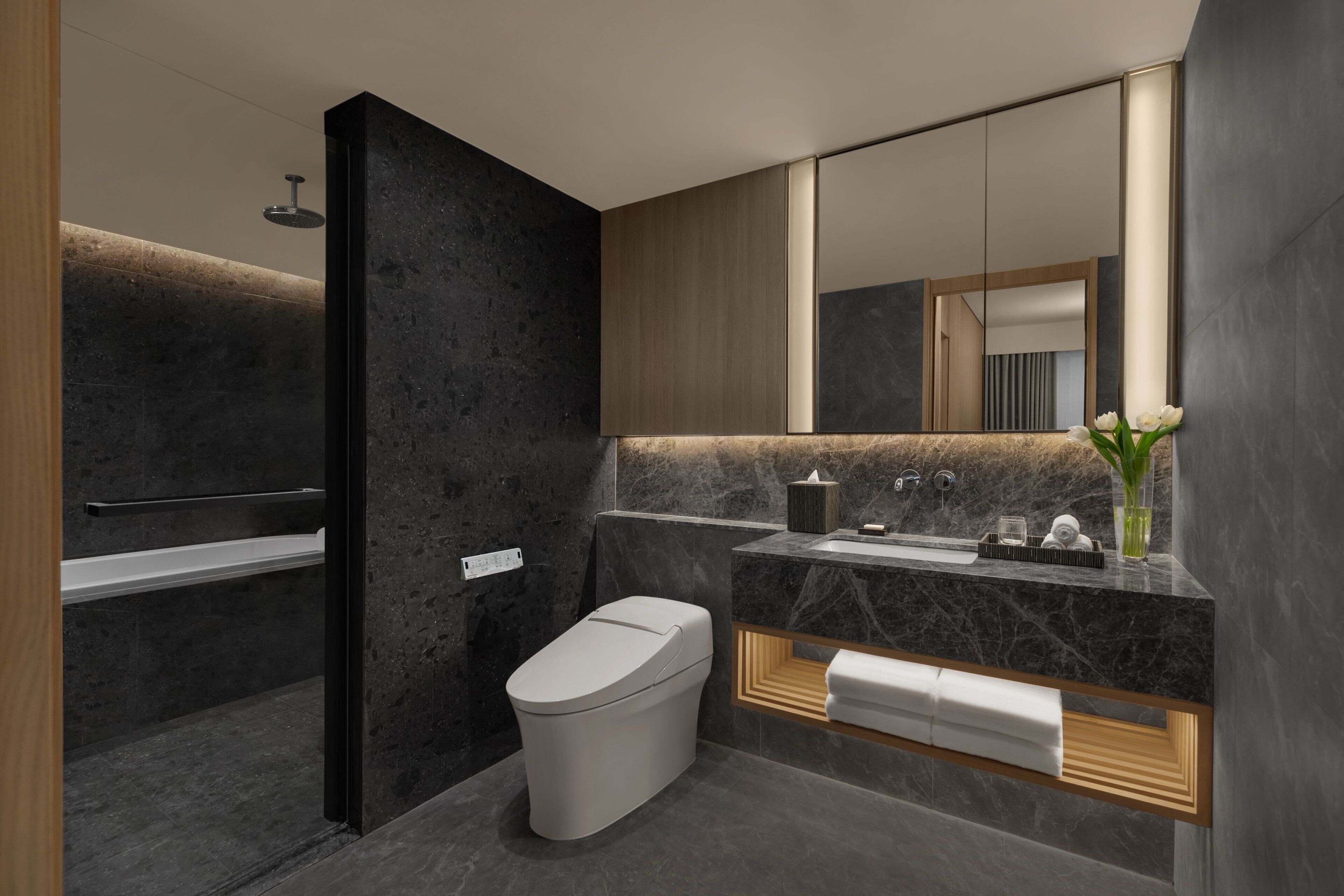 A modern bathroom with a dark stone wall, a white toilet, a wooden vanity, and a large mirror.