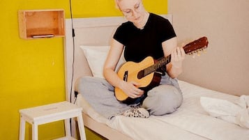Iemand zit op een bed, met een gitaar, een lamp en een klein houten doosje aan de muur.
