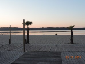 Eine Holzstege führt zu einem Strand mit Palmen und ruhigem Wasser im Hintergrund.