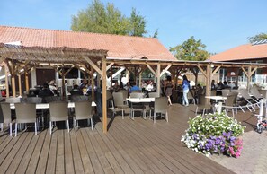 Ein Essbereich im Freien mit Holztischen und -stühlen, der von einer Pergola überdacht wird.