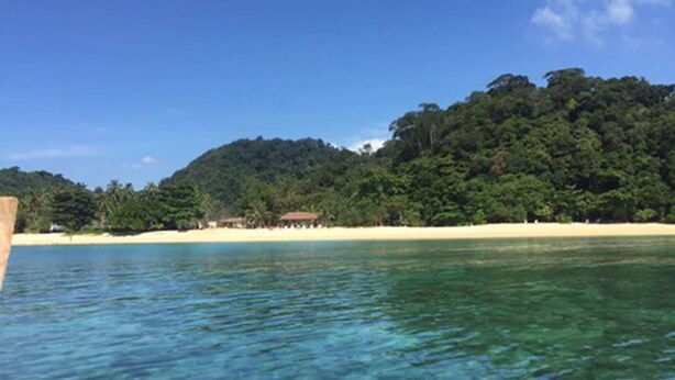 A beach with clear turquoise water, a sandy shore, and a forested hill in the background.