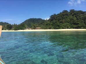A beach with clear turquoise water, a sandy shore, and a forested hill in the background.