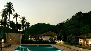 A swimming pool with a wooden deck, surrounded by palm trees and a building with a tiled roof.
