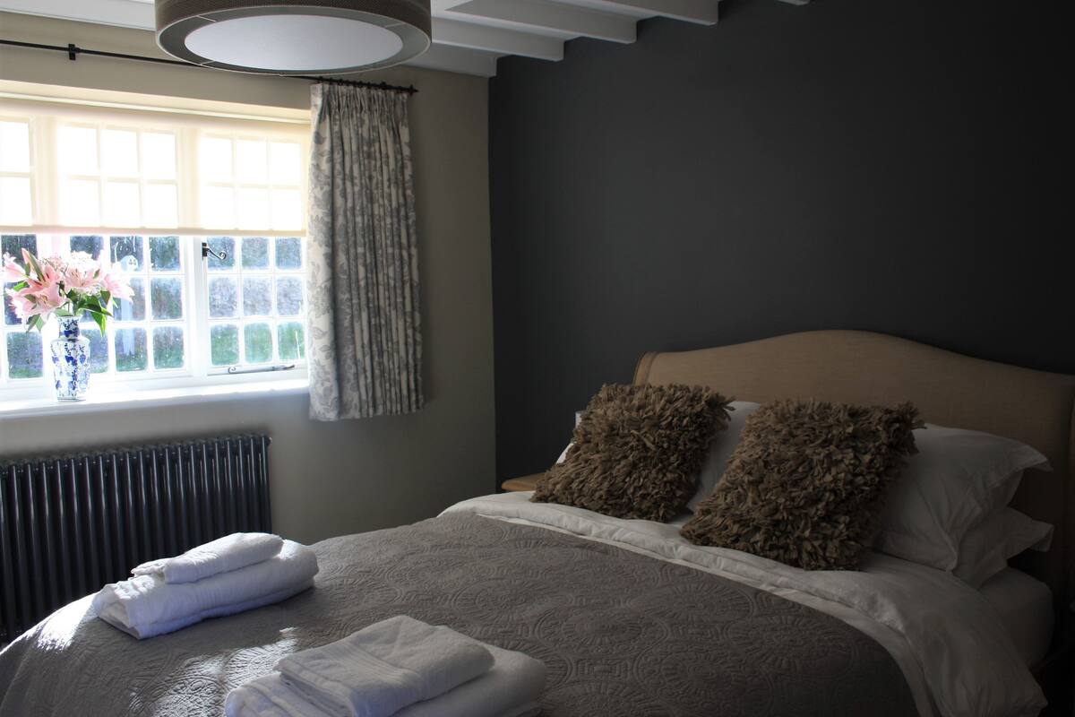 A bedroom with a bed, pillows, a window with curtains, and a vase with flowers.