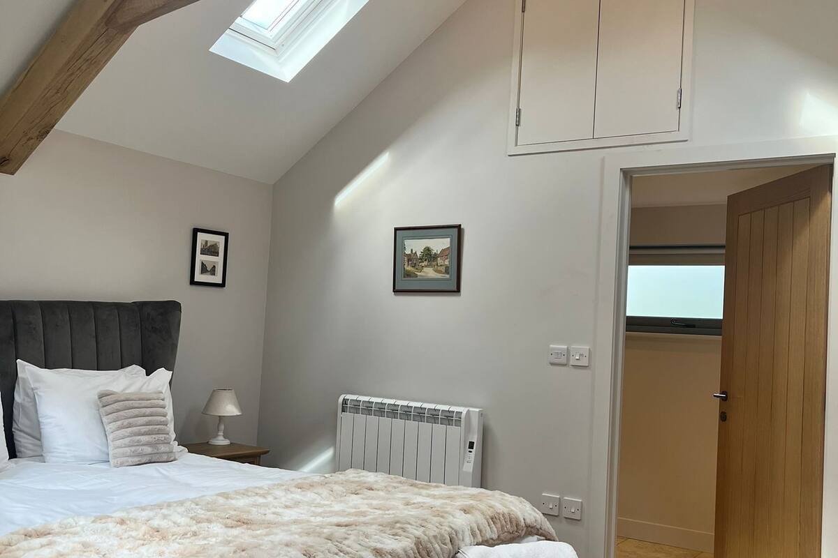 A bedroom with a sloped ceiling, a bed with a fluffy blanket, a bench with towels, a radiator, and a chandelier.