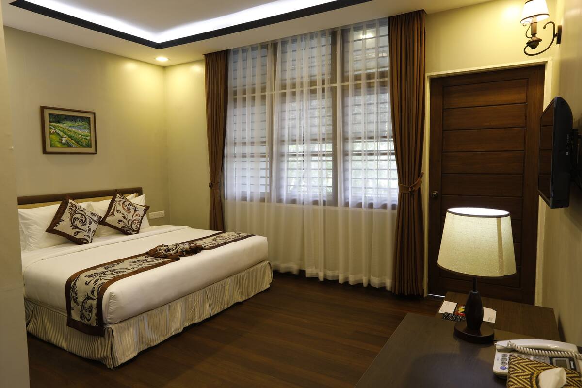 Une chambre d’hôtel avec un grand lit, une table de chevet avec une lampe, un téléphone et une fenêtre avec des rideaux.
