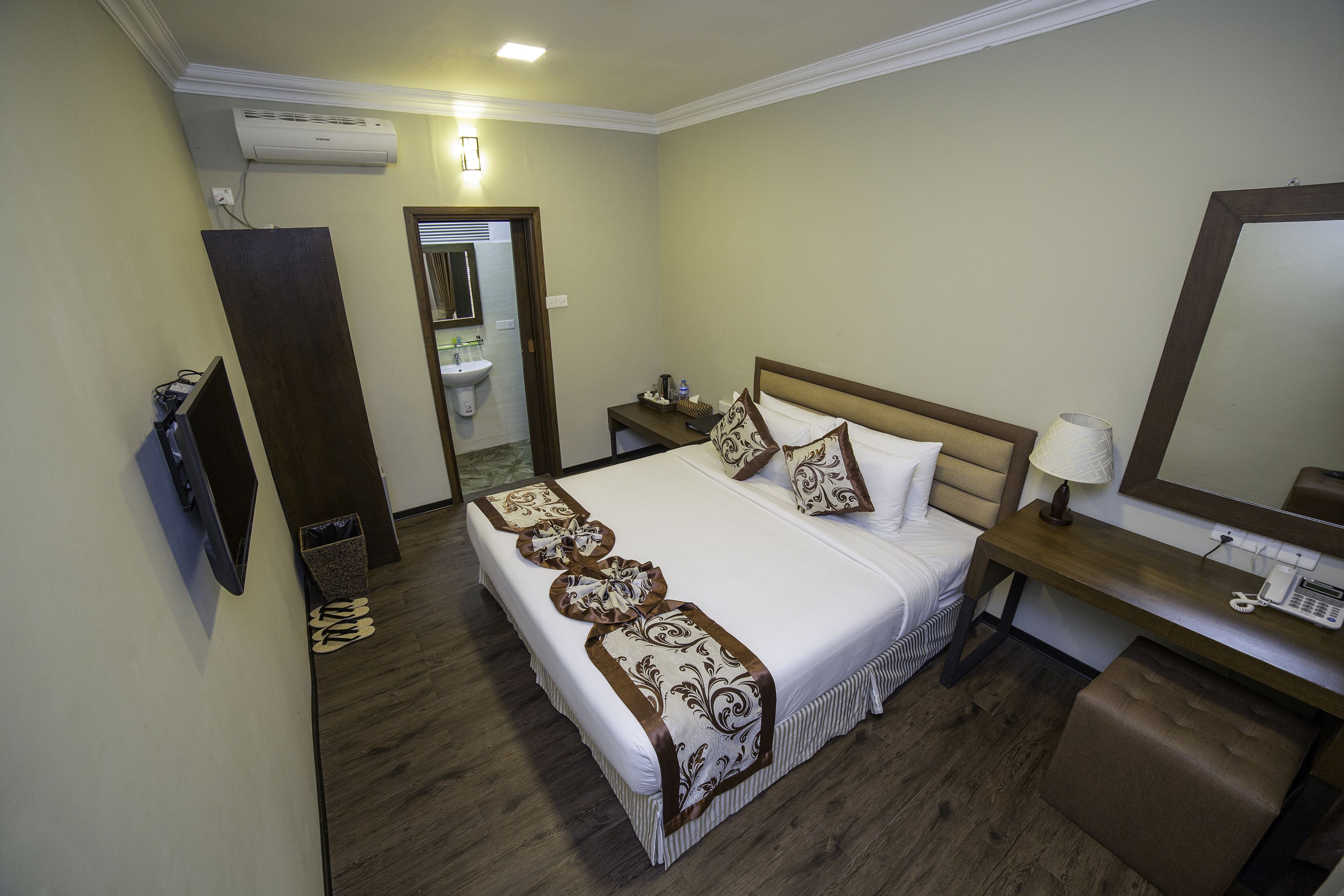 A hotel room with a large bed, bedside tables, a desk with a lamp, a mirror, and an air conditioning unit.