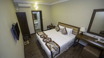 A hotel room with a large bed, bedside tables, a desk with a lamp, a mirror, and an air conditioning unit.