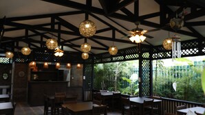 Uma área de refeições interna com mesas e cadeiras de madeira, luminárias esféricas pendentes e vista para o verde através de uma janela com treliça.
