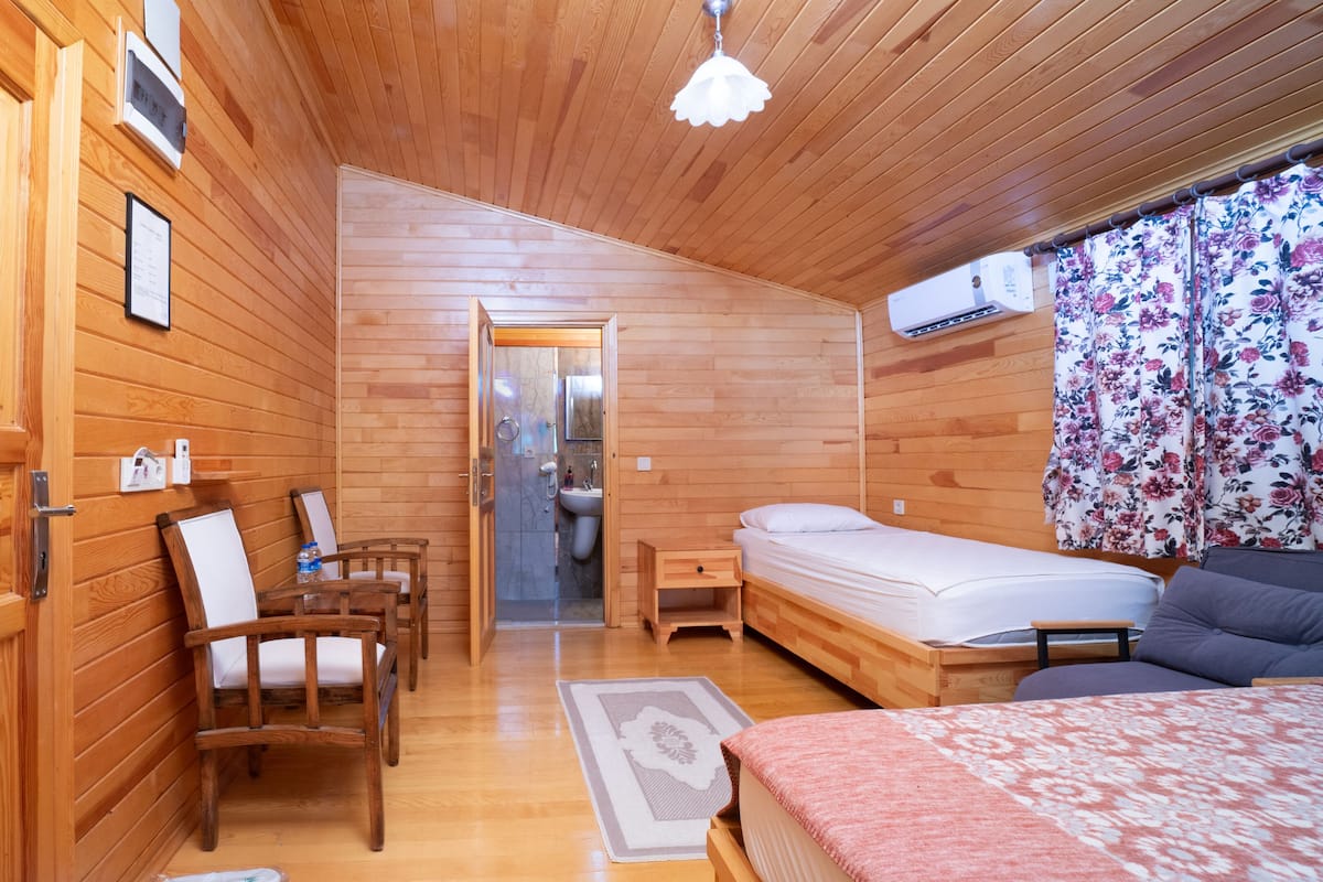 Habitación de cabaña de madera con cama, escritorio, silla, mesita pequeña, ducha y ventana con cortinas florales.