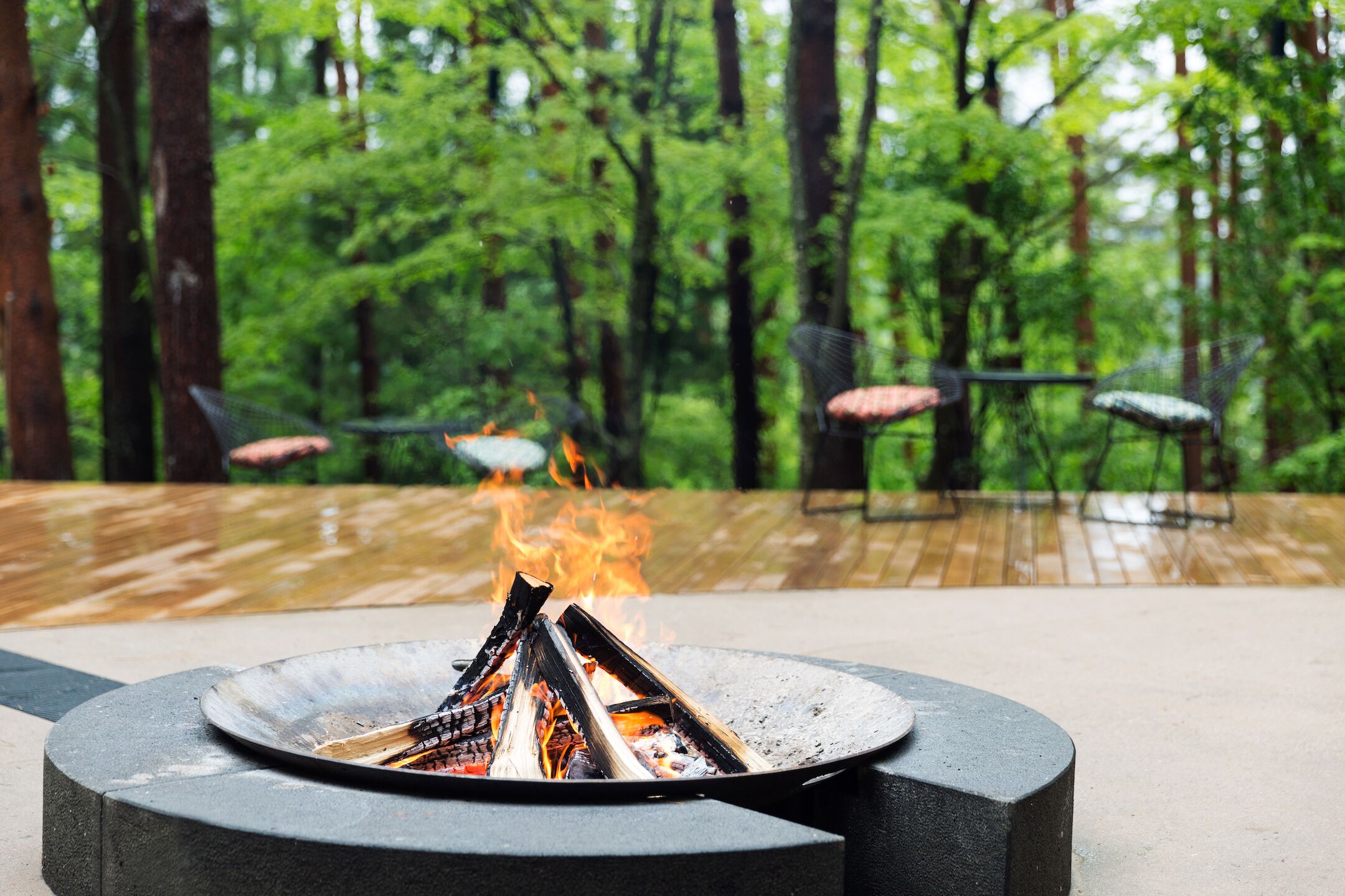 A fire pit with logs in a circular metal basin, surrounded by a wooden deck with chairs.