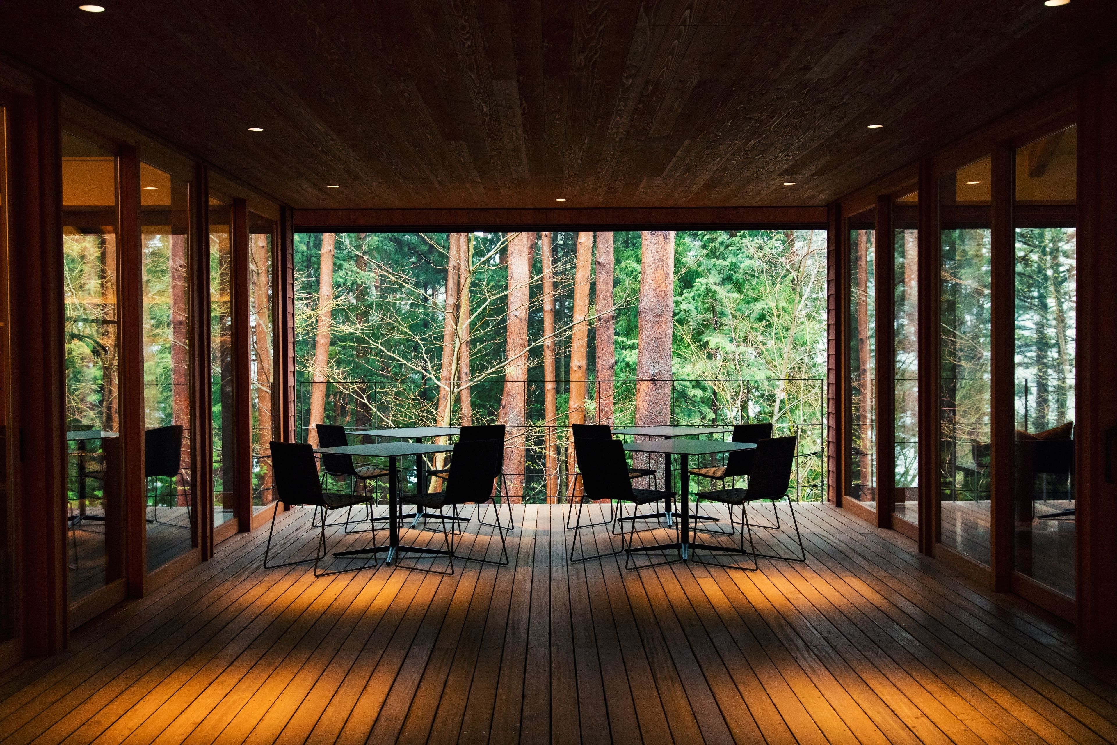 A wooden deck with a table and chairs, large windows, and a forest view.