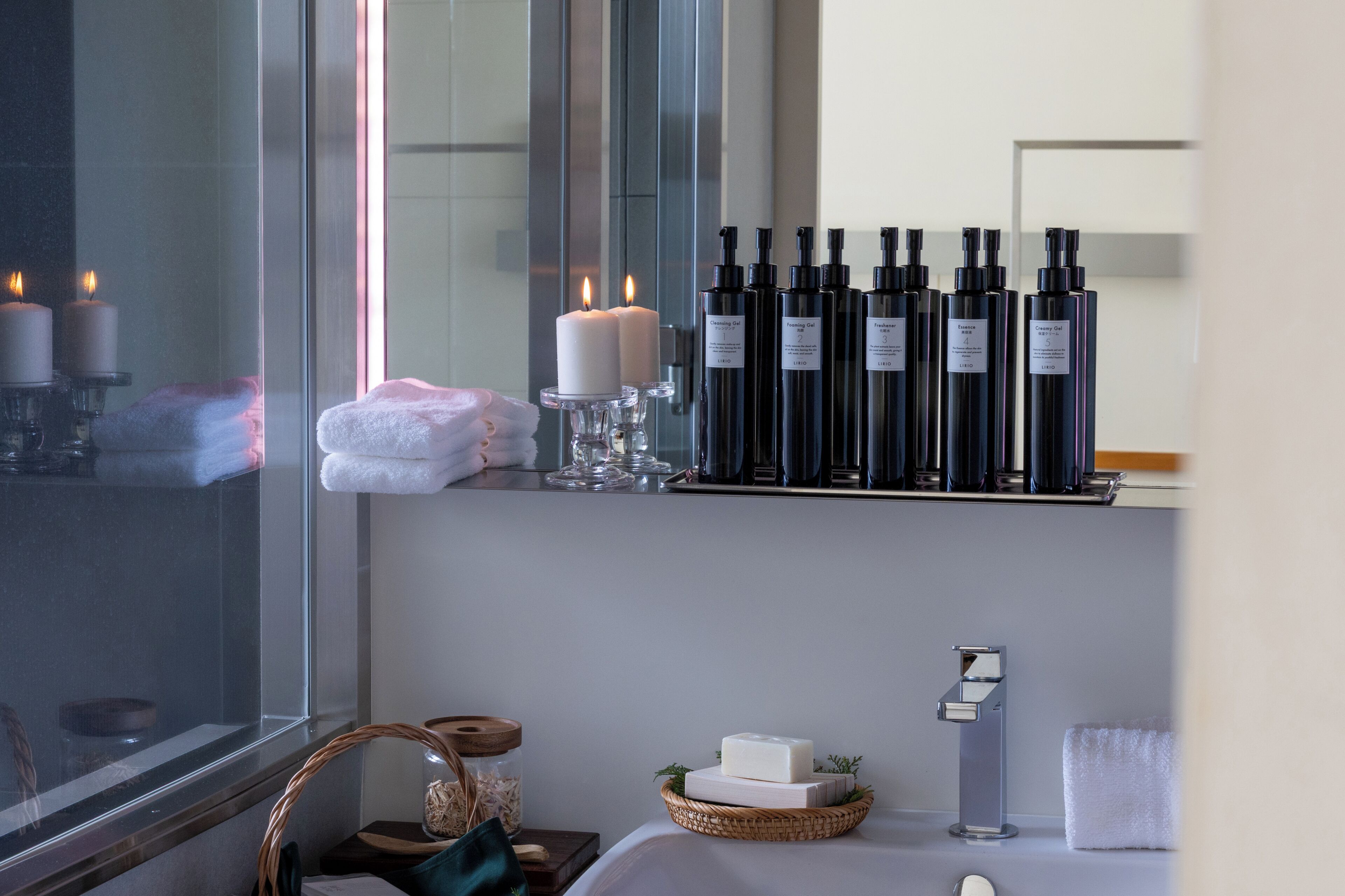 A bathroom with a mirror, candles, bottles of shampoo, a towel, and a basket with a bar of soap.