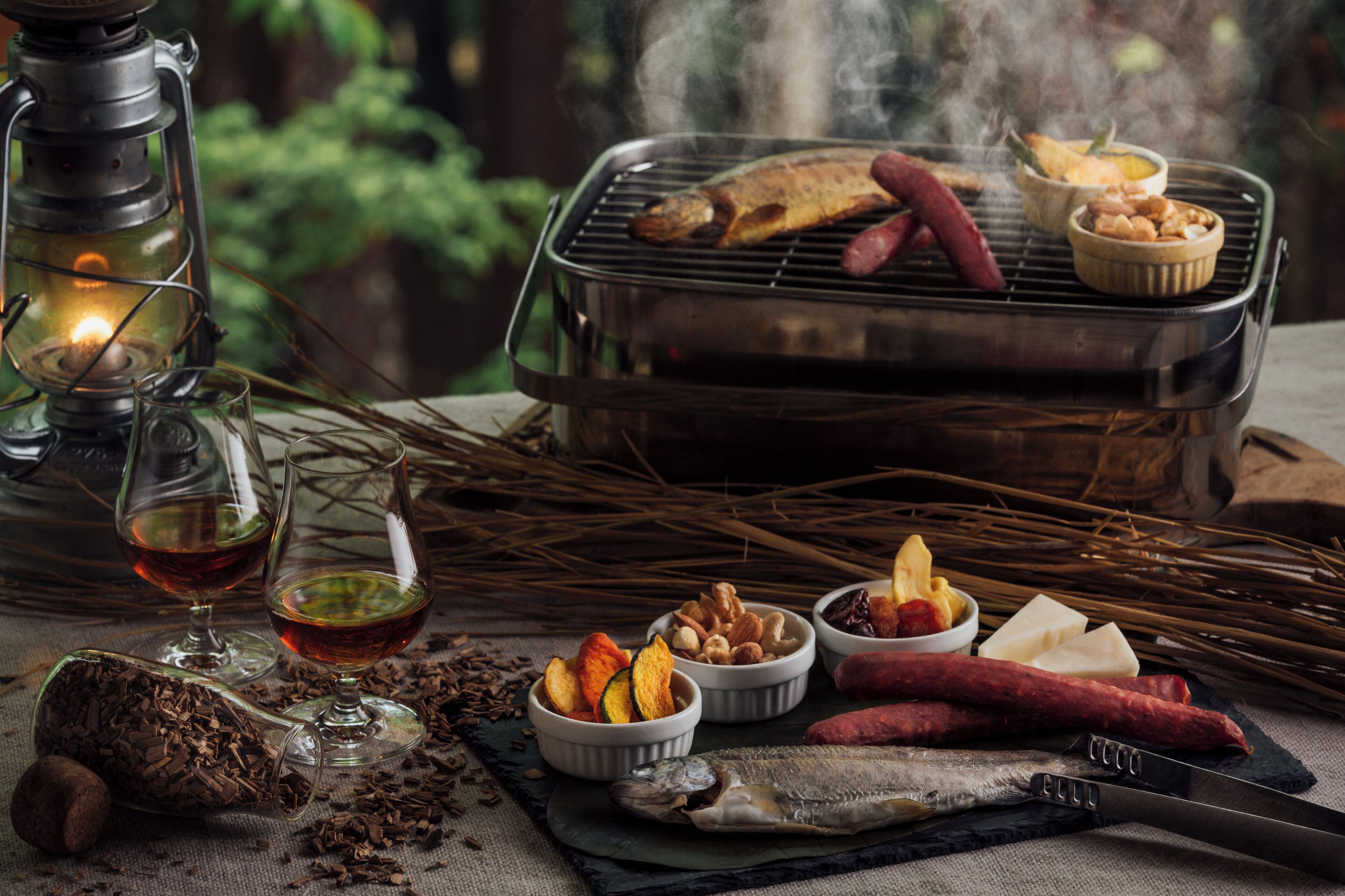 A rustic outdoor dining setup with a grill, food, and a lantern.
