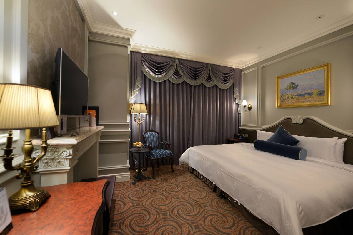 A hotel room with a large bed, a desk with a lamp, a chair, and a painting on the wall.
