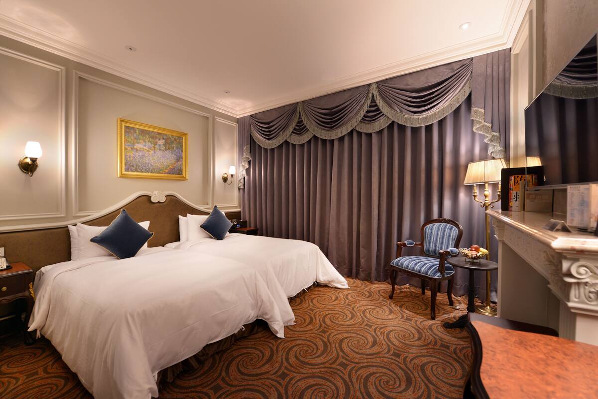 A hotel room with a large bed, two pillows, a nightstand, a chair, a small table, and a painting on the wall.