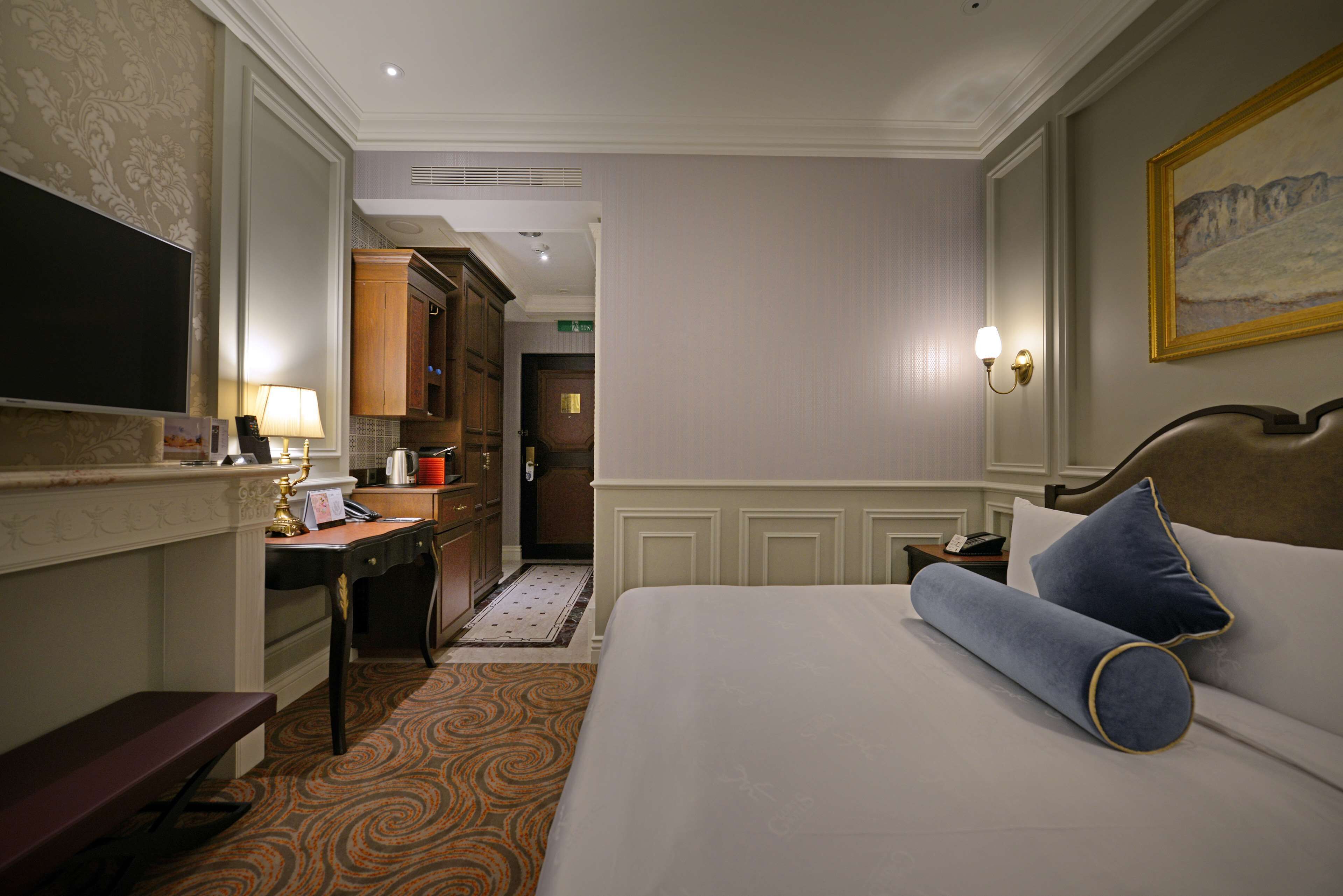 A hotel room with a bed, a desk, a chair, a television, and a painting on the wall.