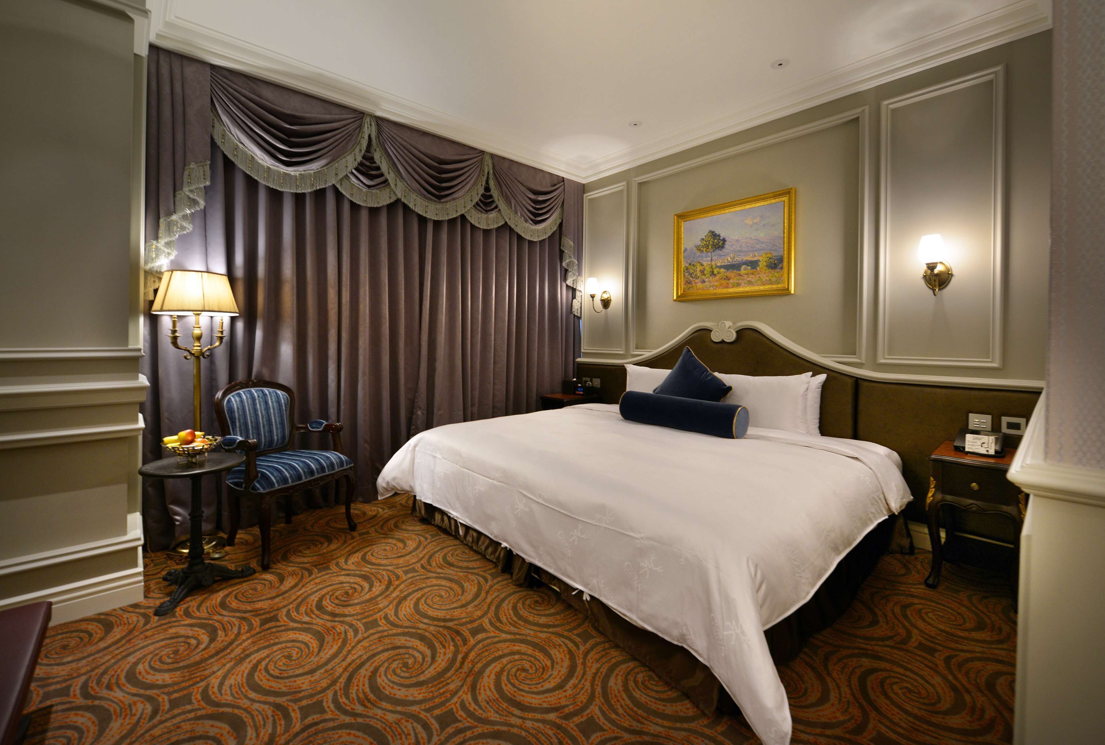 Habitación de hotel con una cama grande, una silla, una mesa con lámpara y un cuadro en la pared.
