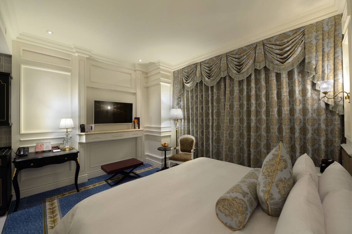 A hotel room with a large bed, a desk with a lamp, a chair, a television, and a patterned curtain.