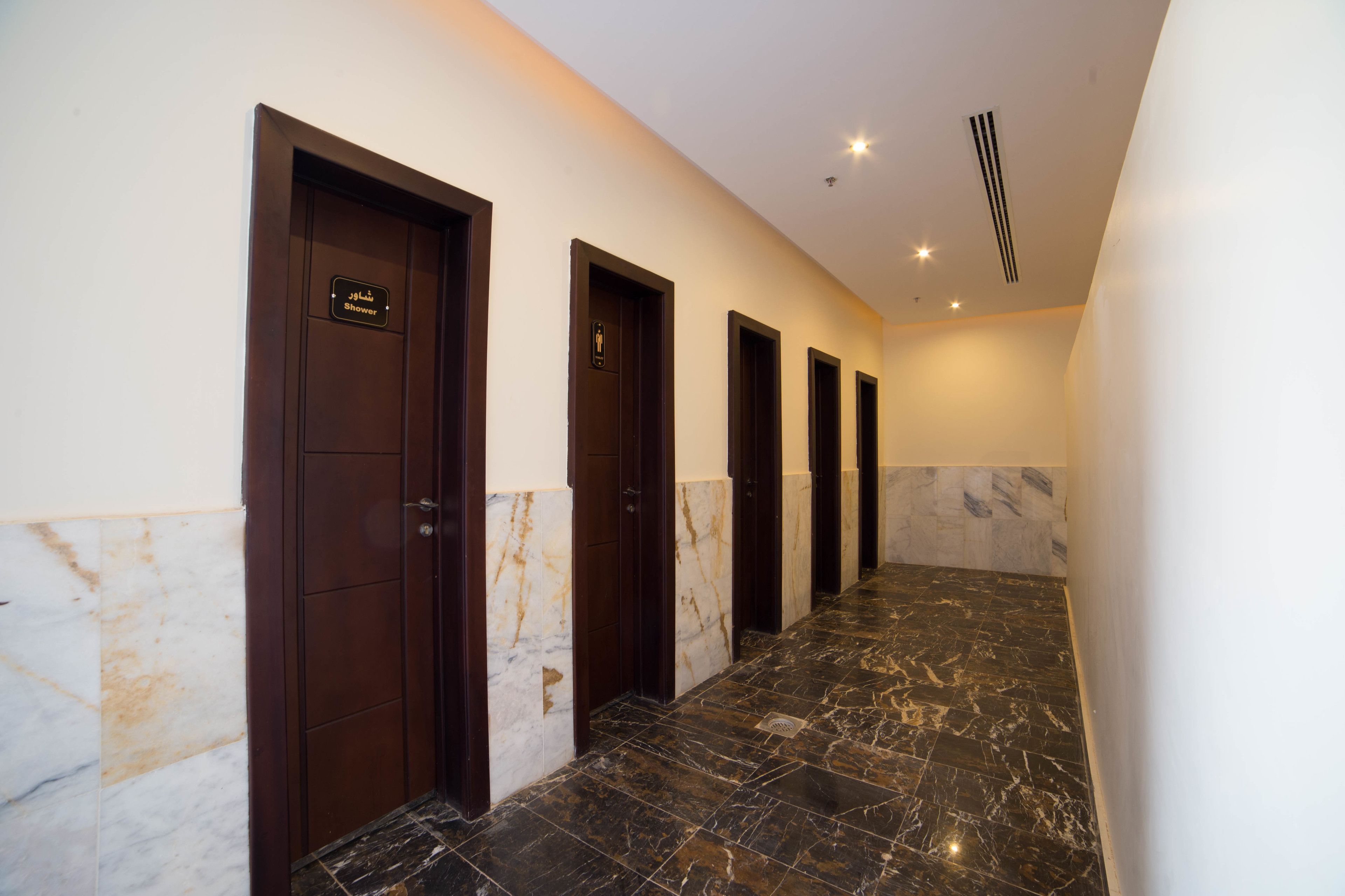 Un couloir d’hôtel avec un sol en marbre et des portes en bois foncé, dont certaines sont signalées comme donnant accès à des douches.