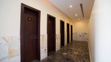 Un couloir d’hôtel avec un sol en marbre et des portes en bois foncé, dont certaines sont signalées comme donnant accès à des douches.