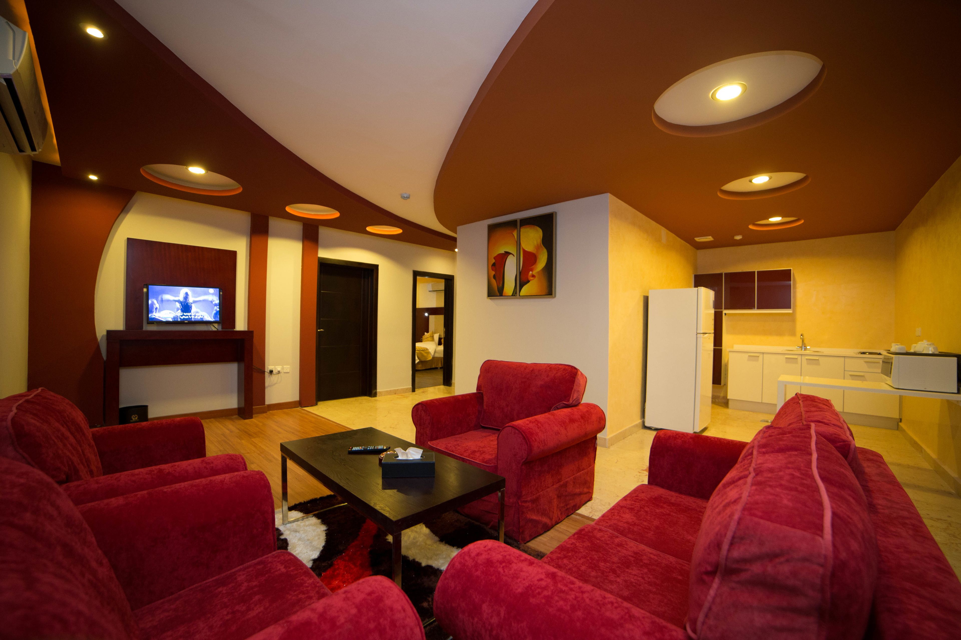 A modern living room with red sofas, a black coffee table, and a flat-screen TV. There is a kitchen area with white cabinets and a refrigerator.