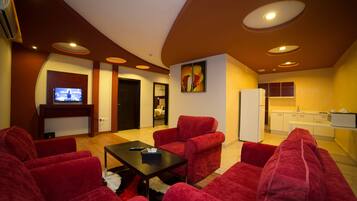 A modern living room with red sofas, a black coffee table, and a flat-screen TV. There is a kitchen area with white cabinets and a refrigerator.
