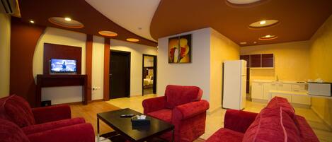 A modern living room with red sofas, a black coffee table, and a flat-screen TV. There is a kitchen area with white cabinets and a refrigerator.