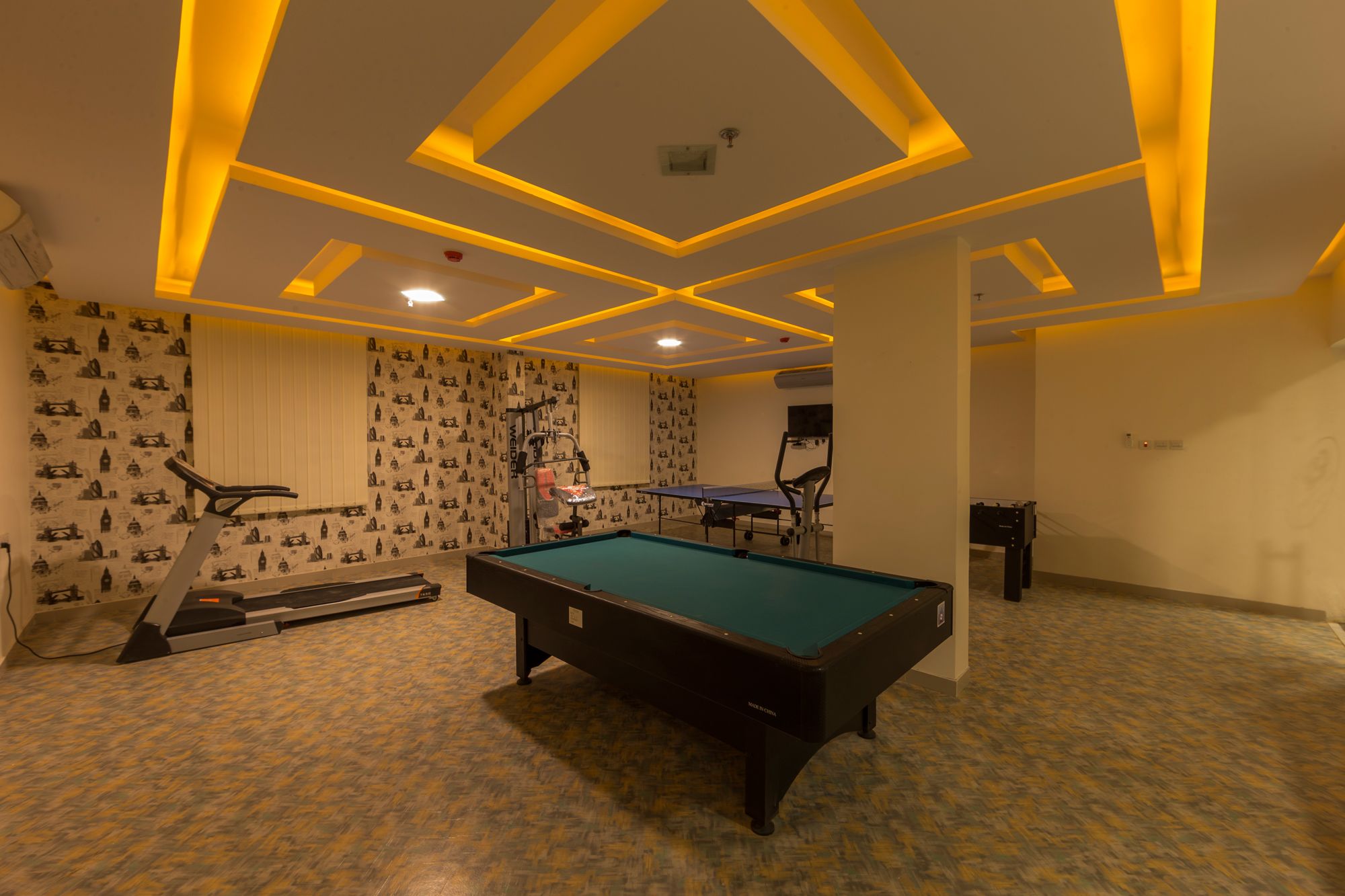 A pool table in a room with patterned wallpaper, a treadmill, and a foosball table.