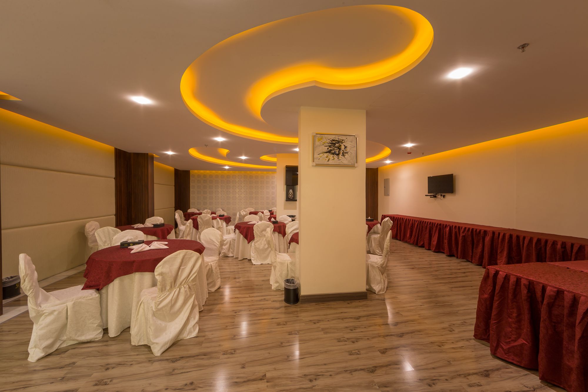 Una sala de banquets amb taules rodones cobertes amb mantells vermells, cadires blanques i un pilar central amb una obra d'art emmarcada.