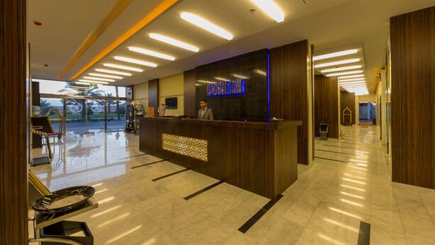 Un espace d’accueil d’hôtel au design moderne, comprenant un comptoir d’accueil, un panneau numérique affichant le nom de l’hôtel et une porte vitrée donnant sur l’extérieur.
