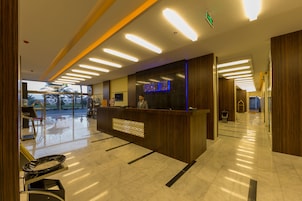 A hotel reception area with a modern design, featuring a reception desk, a digital sign displaying the hotel name, and a glass door leading outside.