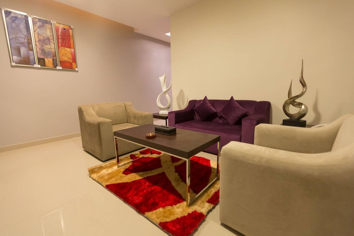 A modern living room with a beige sofa, a purple sofa, and a white armchair. There is a coffee table with a remote control and a decorative item. A red and beige rug is on the floor.