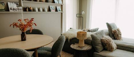 A modern living room with a round dining table, grey upholstered sofa, and a bookshelf with various items.