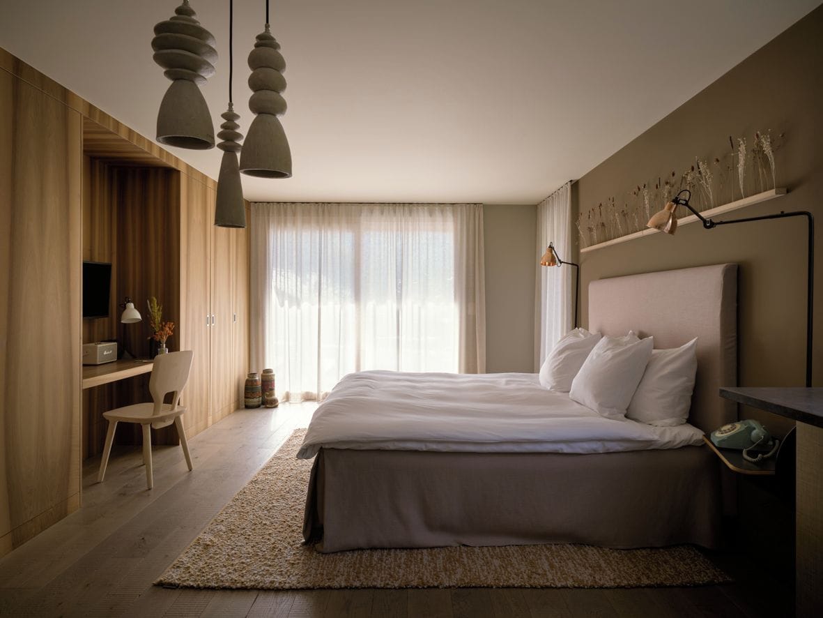 A modern bedroom with a large bed, a desk, and a chair. There is a window with curtains and a wall-mounted shelf with decorative items.